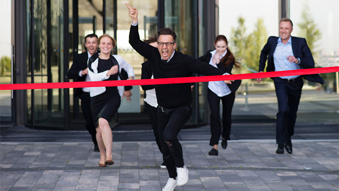 Man in business attire breaks through a red finish line ribbon as colleagues cheer and run behind him, symbolizing success.